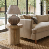 A modern living room features a beige sofa with decorative pillows. A LuxenHome dark oak finish fluted round side table holds a textured lamp. Large windows show an outdoor view, while a woven rug covers the light wood floor, and pillows add accents to the neutral decor.