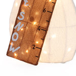 A brown wooden snow measuring stick with white letters spelling SNOW and measurement markings, decorated with small white lights, makes a charming lighted display and yard holiday decor, leaning against a white surface.