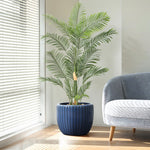 Tall potted palm plant with feathery leaves in a dark blue ceramic flowerpot, featuring a three-dimensional vertical pattern, beside a grey upholstered chair near white blinds in a sunlit room.