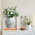 A white and blue Floral Ceramic Vase filled with white flowers and greenery sits on books, charming home décor.