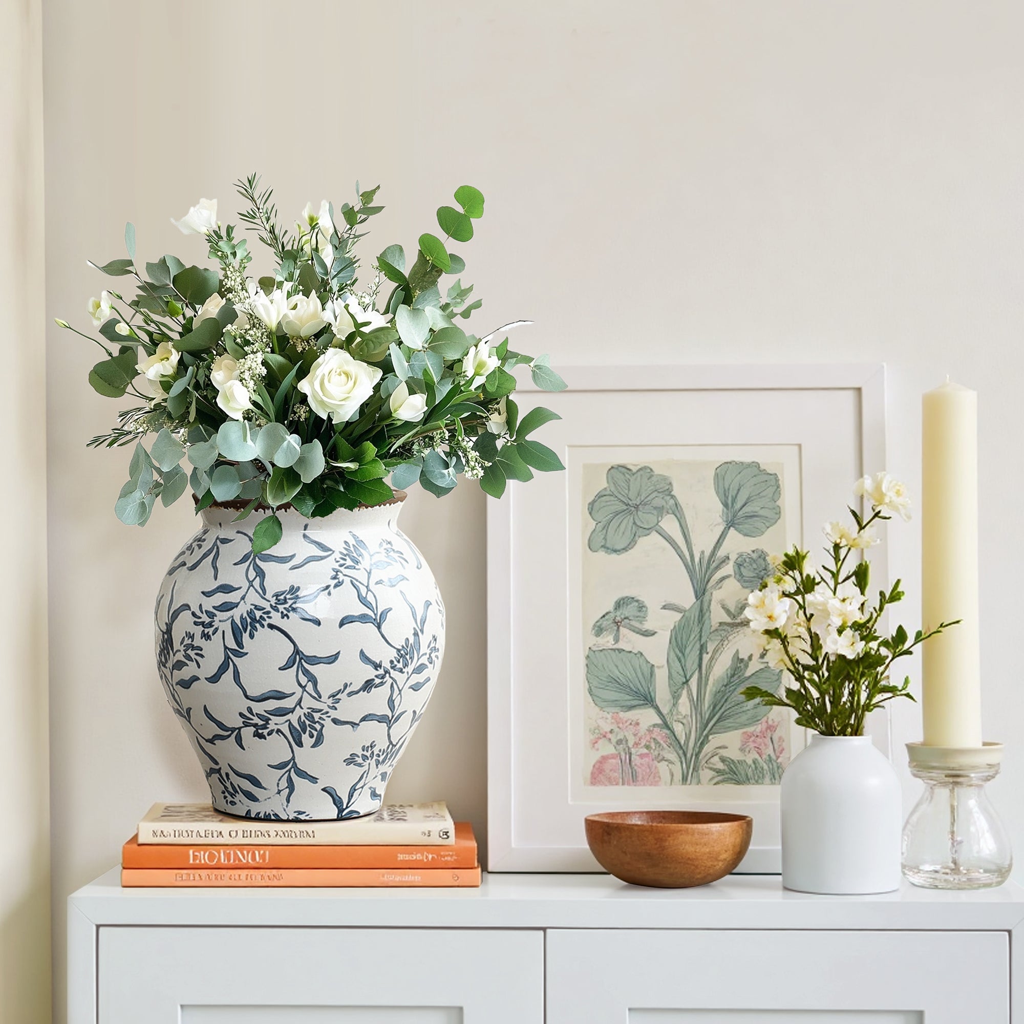 A white and blue Floral Ceramic Vase filled with white flowers and greenery sits on books, charming home décor.