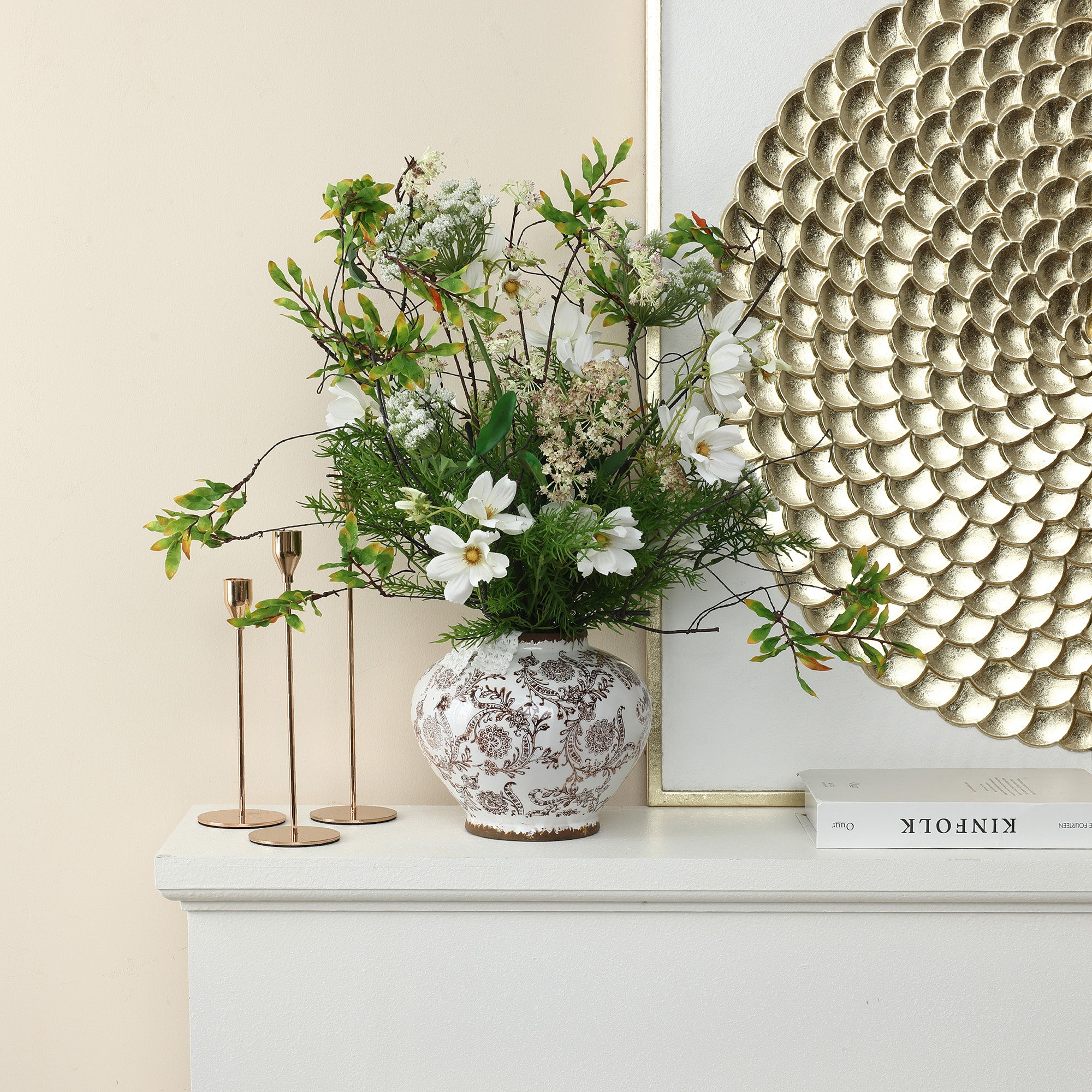 A floral arrangement in an elegant vase sits on a white surface beside gold candlesticks, a book.