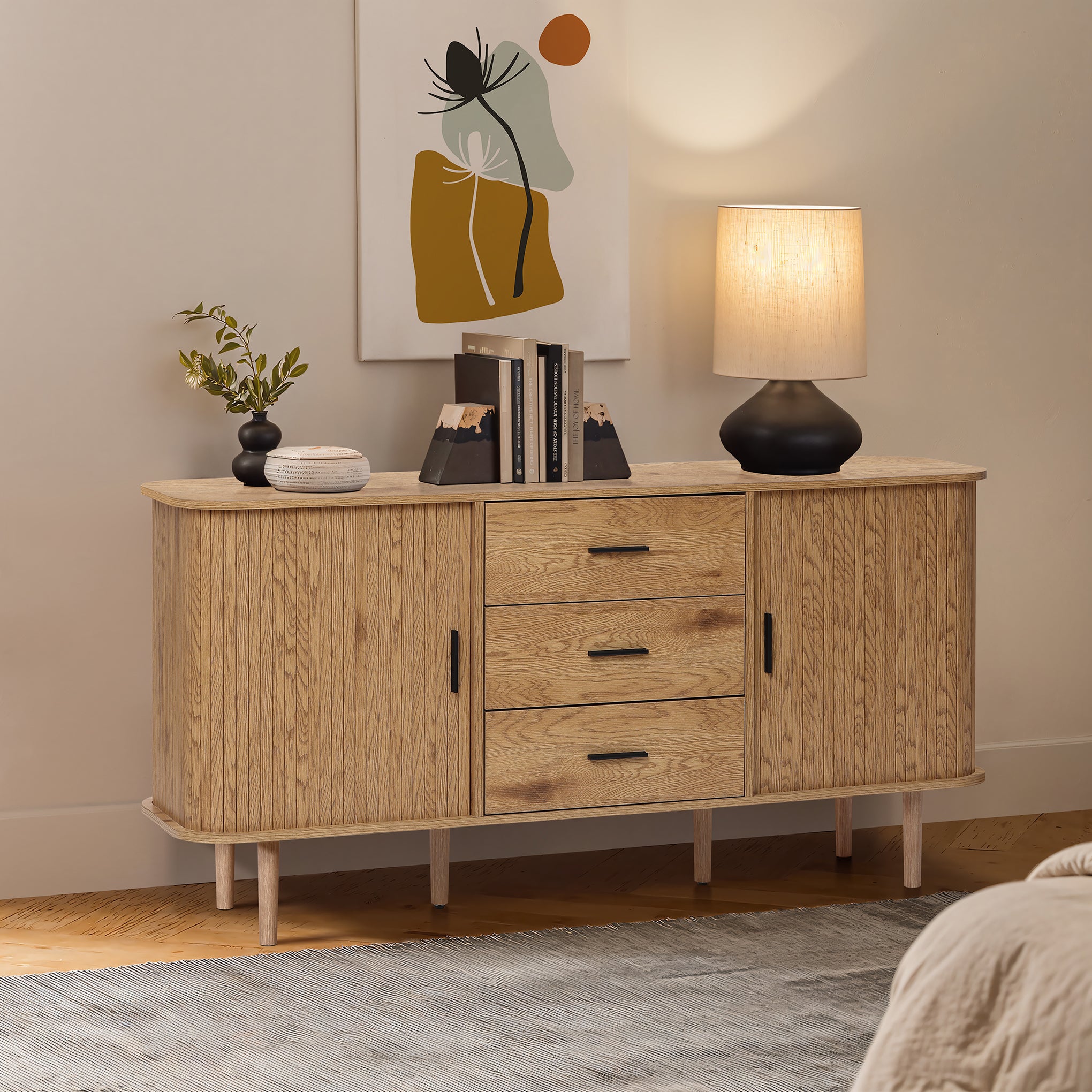 Buffet cabinet sideboard, a mid century wood storage unit with three drawers and two cabinets, topped with books, a lamp, a plant, and décor, standing against a wall with abstract art.