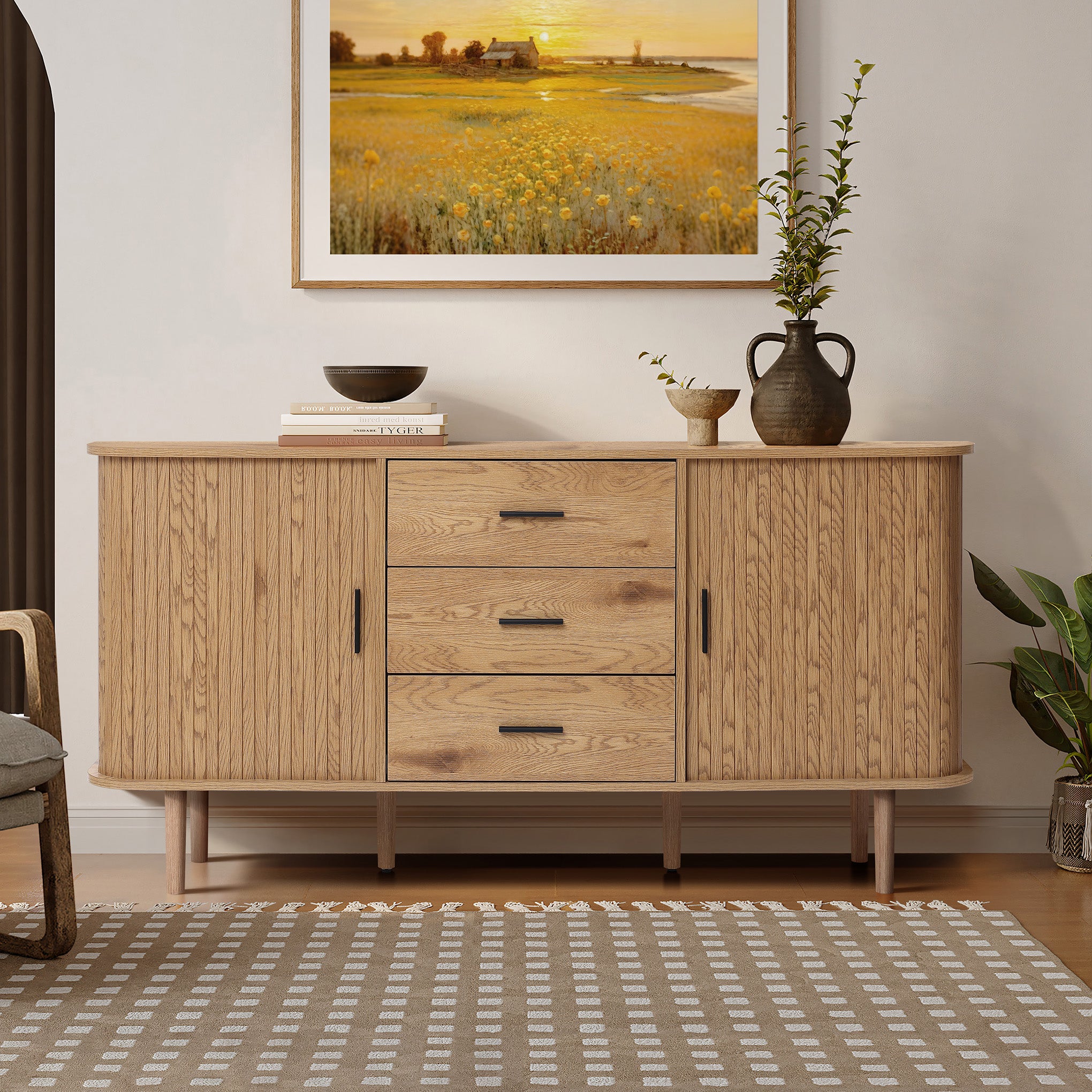 Buffet cabinet sideboard, a mid century wood storage cabinet with three drawers and two cabinets, topped with books, vases, and decor, standing against a wall with a framed landscape painting.