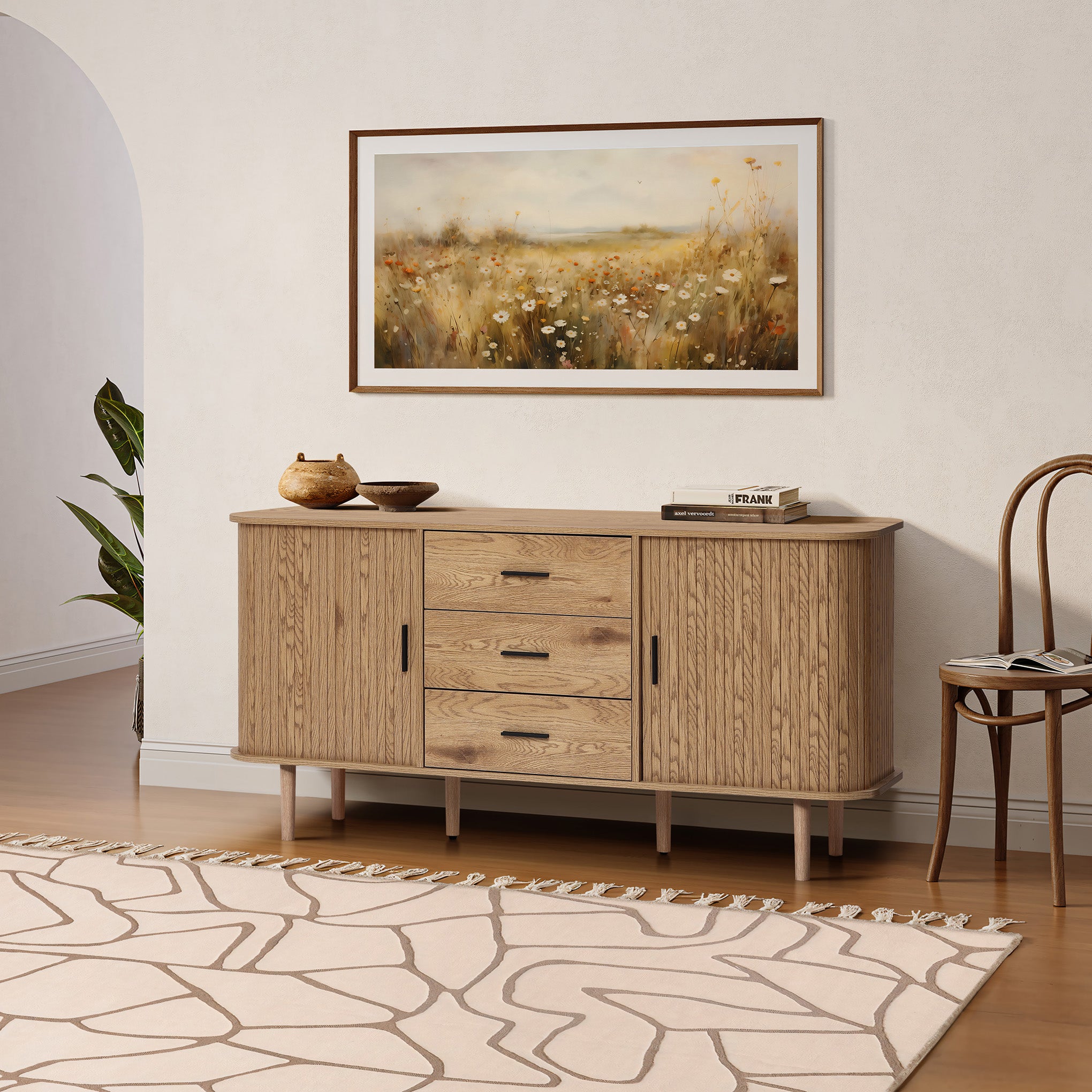 Buffet cabinet sideboard, a mid century storage cabinet with three drawers and two cabinets, placed against a white wall below a framed landscape painting, with a rug, chair, and potted plant completing the room scene.