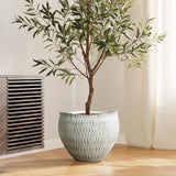 A tall potted olive tree with thin green leaves stands on a wooden floor near a vent, placed in a LuxenHome White textured resin round planter, large, featuring a geometric pattern. Soft natural light filters through a sheer curtain.