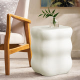 A small curvy side table crafted from modern MgO material sits elegantly with a clear vase and green leaves beside a light wooden armchair adorned with a gray cushion.