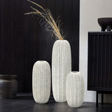 Three white ceramic vases of varying heights stand against a black wall. The LuxenHome Modern geometric pattern resin tall vase, 25 h, contains long dried grasses. Nearby is an antique effect vase. A black cabinet and another dark vase are visible in the background on a tiled floor.