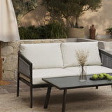 The outdoor patio features a light-colored sofa and an elegant black table with a decorative vase and green apples. A chic outdoor loveseat crafted from PE rattan furniture adds comfort, while the stone wall and lush plants create a serene backdrop.