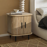 The LuxenHome Aria oak finish nightstand, featuring a slatted sliding door and sleek black handles, stands by the bed. Books rest on top beside a bowl and sculptural vase. A patterned rug peeks from beneath, adding charm to the cozy mid-century modern setting.