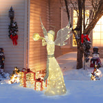 Pre-lit angel stands in the snow beside gift boxes outside a house adorned with festive wreaths.