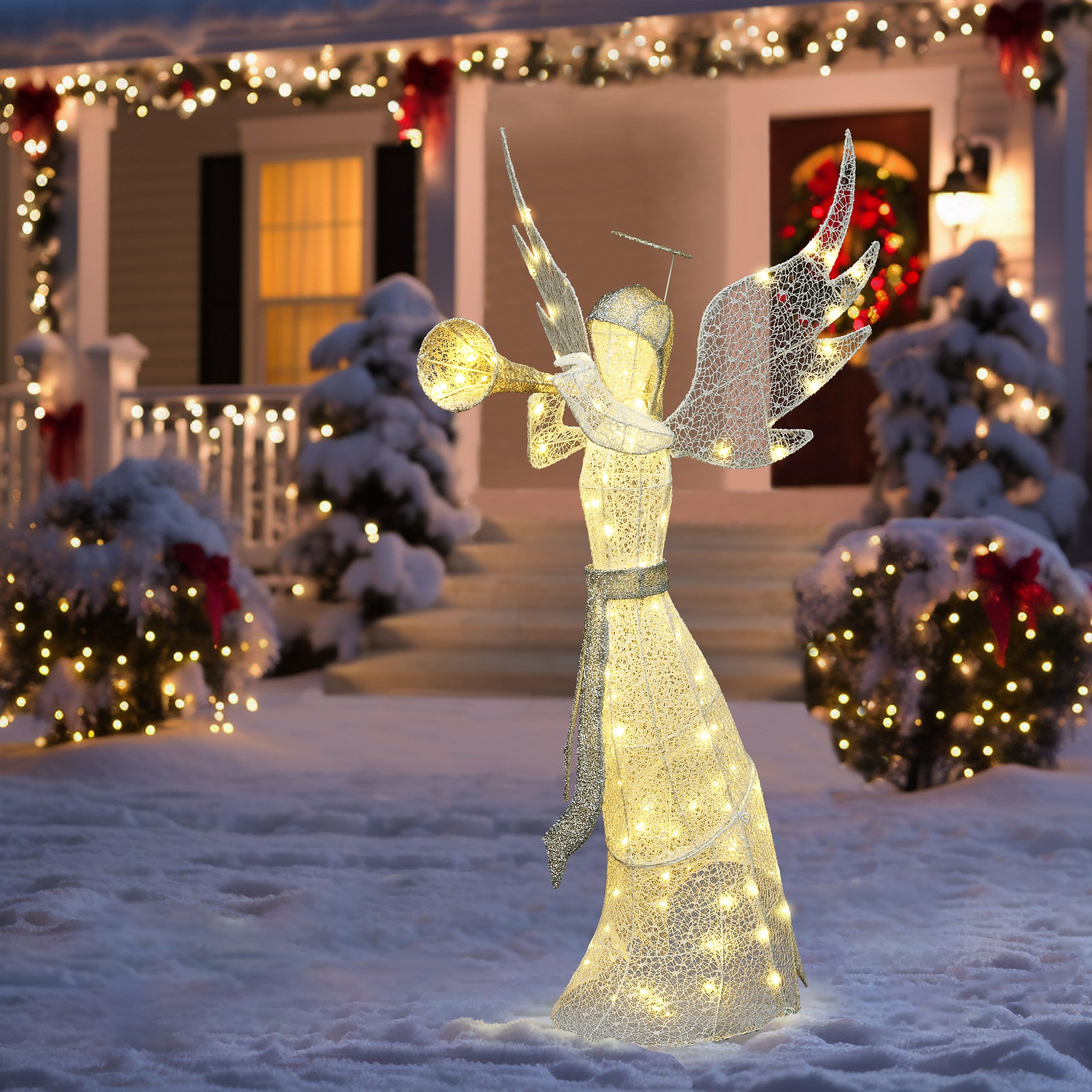 Pre-lit angel stands with trumpet in a snowy yard, a house adorned in christmas lights.