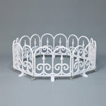 Small white circular fence with decor scroll ,pointed designs, against plain gray background.