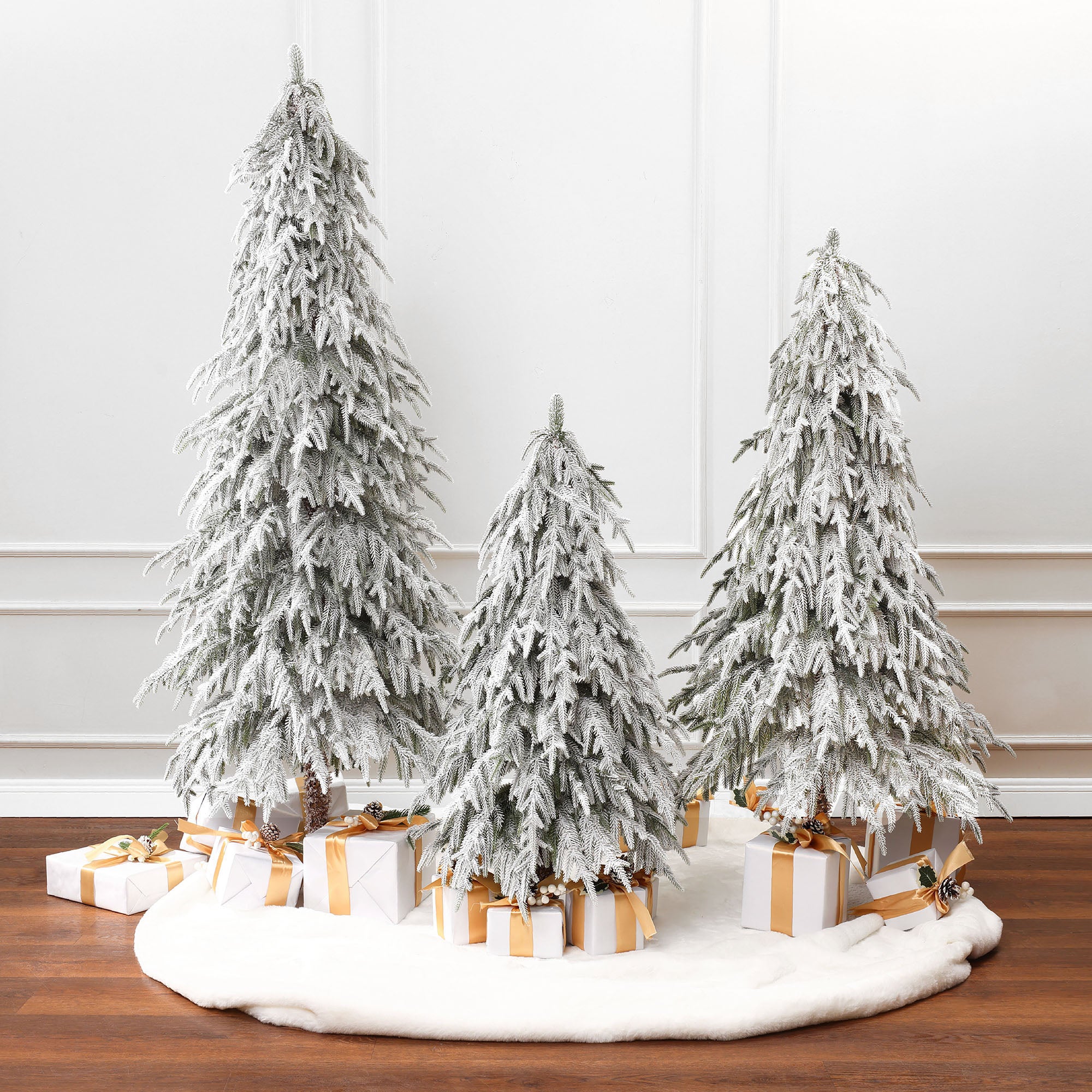 3 snow-flocked cedar trees are arranged on white circular rug with wrapped gifts in white ,gold underneath, againstwhite paneled wall.