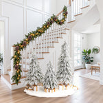 Staircase decor with pine garland, lights sits beside 3 snow-flocked cedar trees with wrapped presents on white mat.