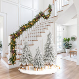 Staircase decor with pine garland, lights sits beside 3 snow-flocked cedar trees with wrapped presents on white mat.