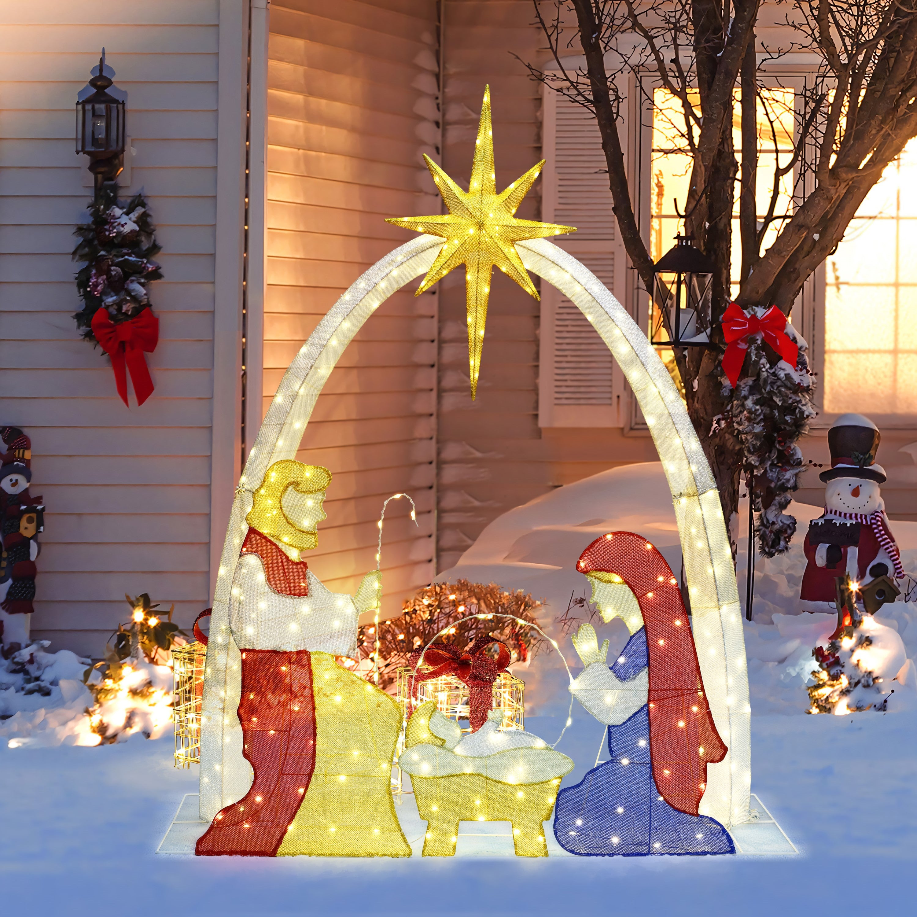 Pre-lit nativity display with figures of Mary, Joseph, baby Jesus under a star on snow,  in front of house beautiful decor. 