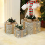 3 silver tinsel gift boxes with bows, pine accents are arranged under Christmas tree on white furry rug.