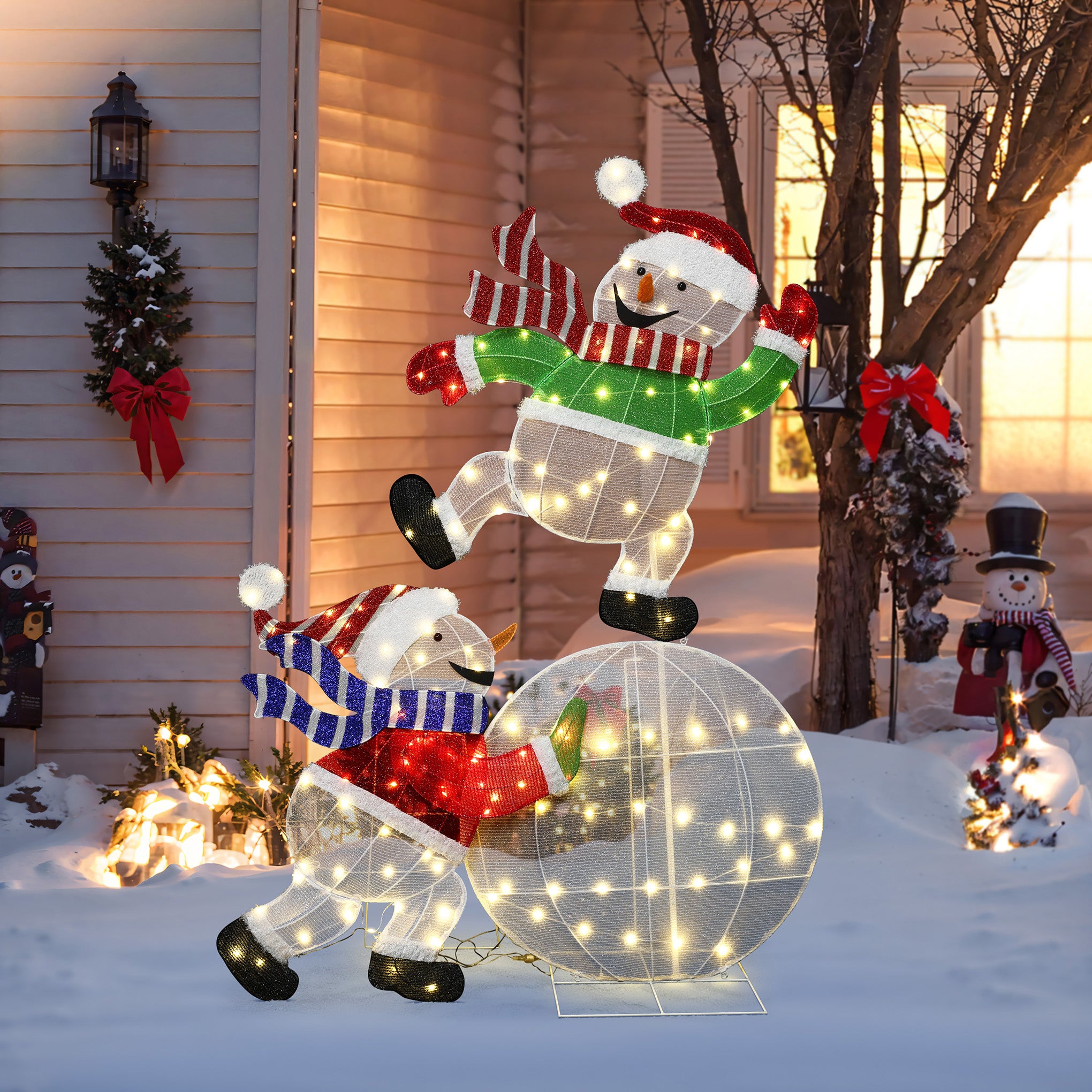 2 illuminated snowman decor, part of an led , stand in snowy yard,one atop glowing snowball, the other pushing.