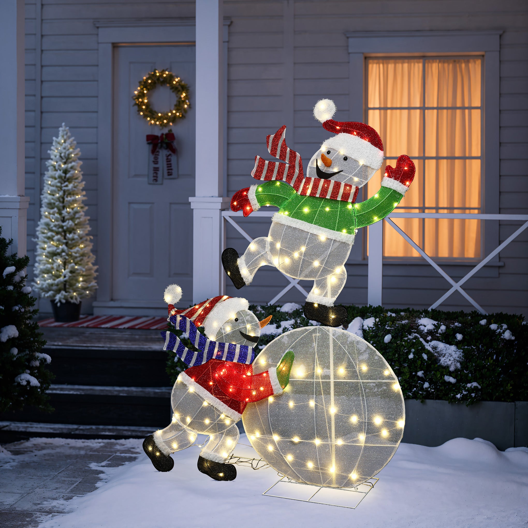 2 illuminated snowman holiday decor ,one climbing ball, the other stand in front of house adorned with lights, wreath, snow.