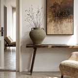 A minimalist interior features a rustic wooden console table with a LuxenHome 14.6 h Rustic Brown Terracotta Cauldron Vase filled with branches. Above, a nature-inspired painting adds charm, while a chair and doorway to another room subtly enrich the scene, completing the homes decor.