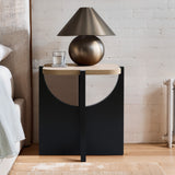 A Bernard black and light wood finish end/side table by LuxenHome stands beside a white brick wall, topped with a sleek metallic lamp and a clear glass of water.