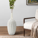 A LuxenHome Modern geometric pattern resin tall trumpet vase stands on a wooden floor next to a checkered armchair with a beige throw. A colorful framed landscape painting hangs on the light gray wall behind, giving an antique touch to the cozy scene.