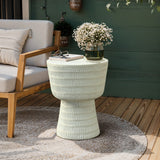 A cozy outdoor patio scene showcases the LuxenHome Meru beige side table, holding a vase of white flowers and glasses. A cushioned wooden chair and textured rug complete the setting against a green wall backdrop.