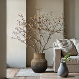 A minimalist living room exudes a vintage vibe with a LuxenHome Brown ombre terracotta round vase, 13.2 h, holding bare branches next to a beige sofa. A woven rug and plant-adorned decor enhance the earthy ambiance on the wooden floor.