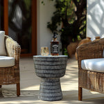 A bottle of whiskey and a glass sit on the LuxenHome Meru outdoor end/side table, black finish, placed between two wicker armchairs with white cushions on a wooden patio. The backdrop is blurred greenery and part of a white building.