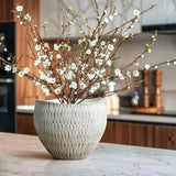 A white textured resin large round planter by LuxenHome sits on a marble countertop in a modern kitchen, with wooden cabinets and blurred kitchen utensils in the background, complementing its sleek design.