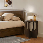 A bedroom scene featuring a wooden bed with beige bedding and a mid-century modern side table. The side table, adorned with two glass bottles and a white lamp, complements the space beautifully. Above the bed hangs a framed abstract artwork, adding an artistic touch to the serene setting.