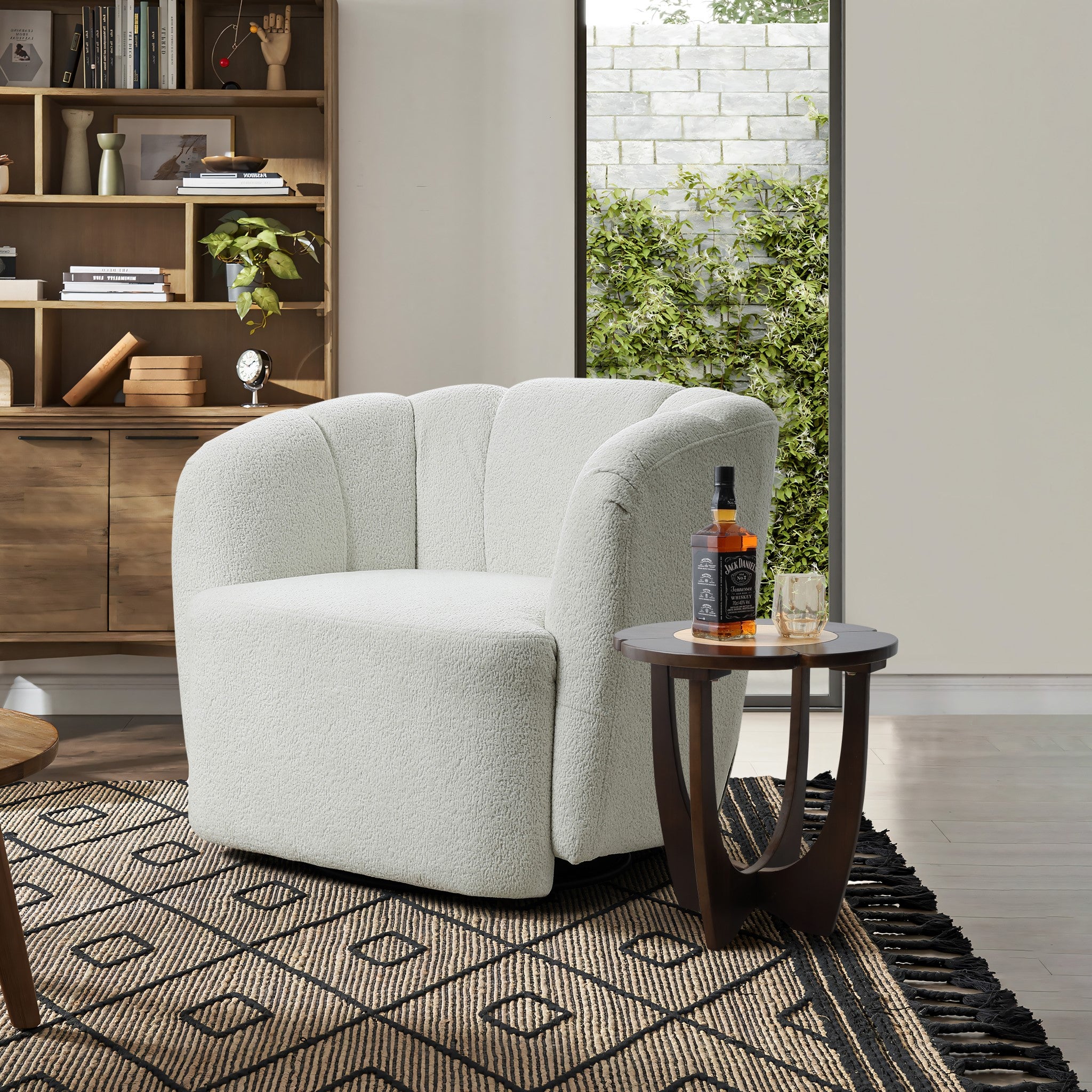 In a cozy living room, a plush armchair sits beside the Sunflower wood end/side table by LuxenHome, holding whiskey and a glass. The modern space features a geometric-patterned rug, natural wood bookshelf, and a window view of greenery.