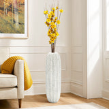 A LuxenHome Modern geometric pattern resin tall vase, 25 h, in white holds yellow flowers with thin branches. Its on a wooden floor beside a cream sofa with a yellow pillow, set against a white wall featuring a colorful painting.