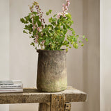 A rustic vase with green and pink flowers graces a wooden table, its charm enhanced by a ginger root brown finish. Beside it, books are stacked invitingly.