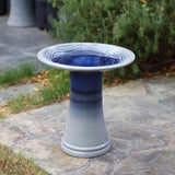 A glazed, two-toned blue and gray ceramic bird bath elegantly stands on a stone patio, surrounded by lush greenery.