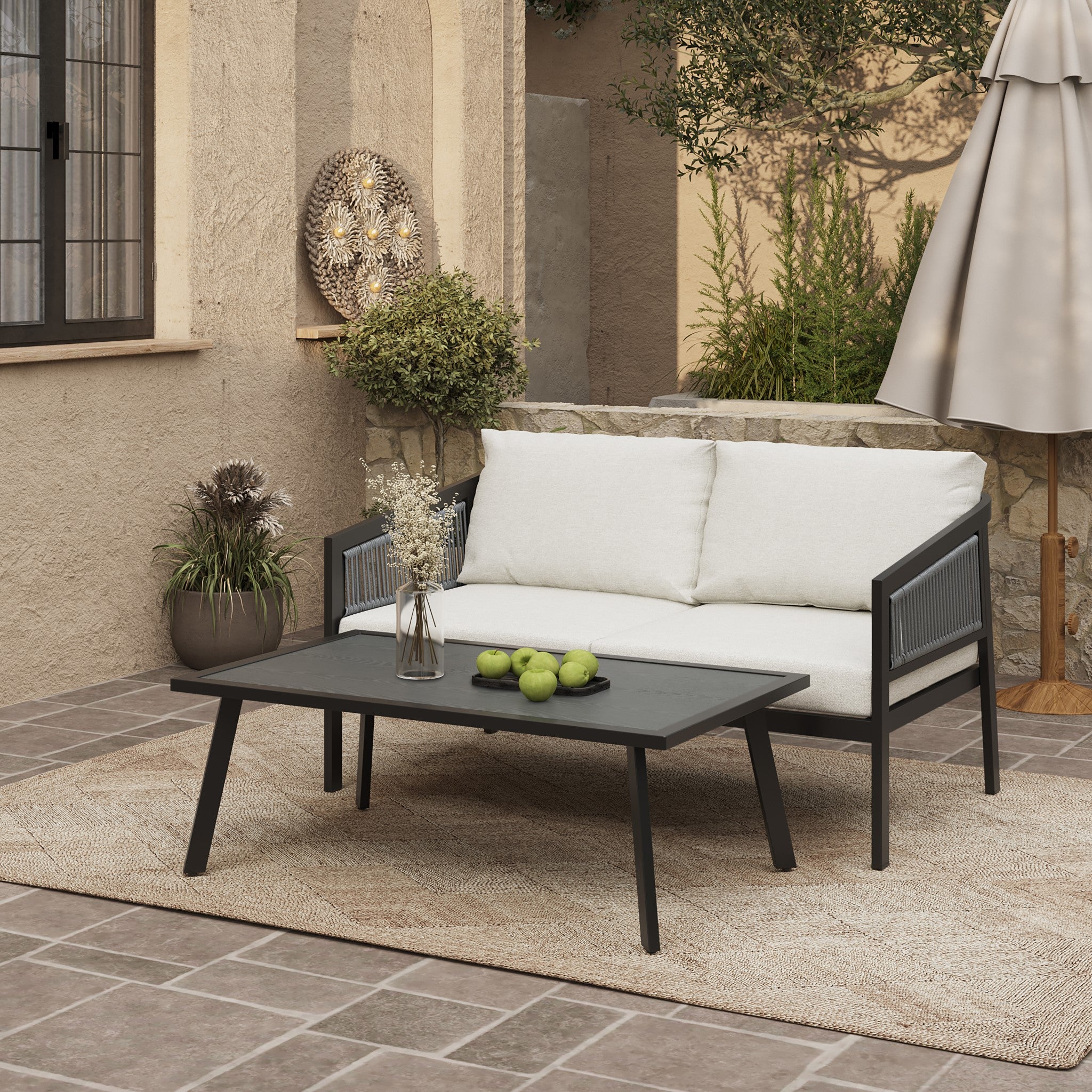 An inviting outdoor patio features a white-cushioned loveseat with a black steel frame, complemented by a black coffee table adorned with green apples. Potted plants and a decorative wall piece add charm, while an umbrella shades the beige rug on the tiled floor.