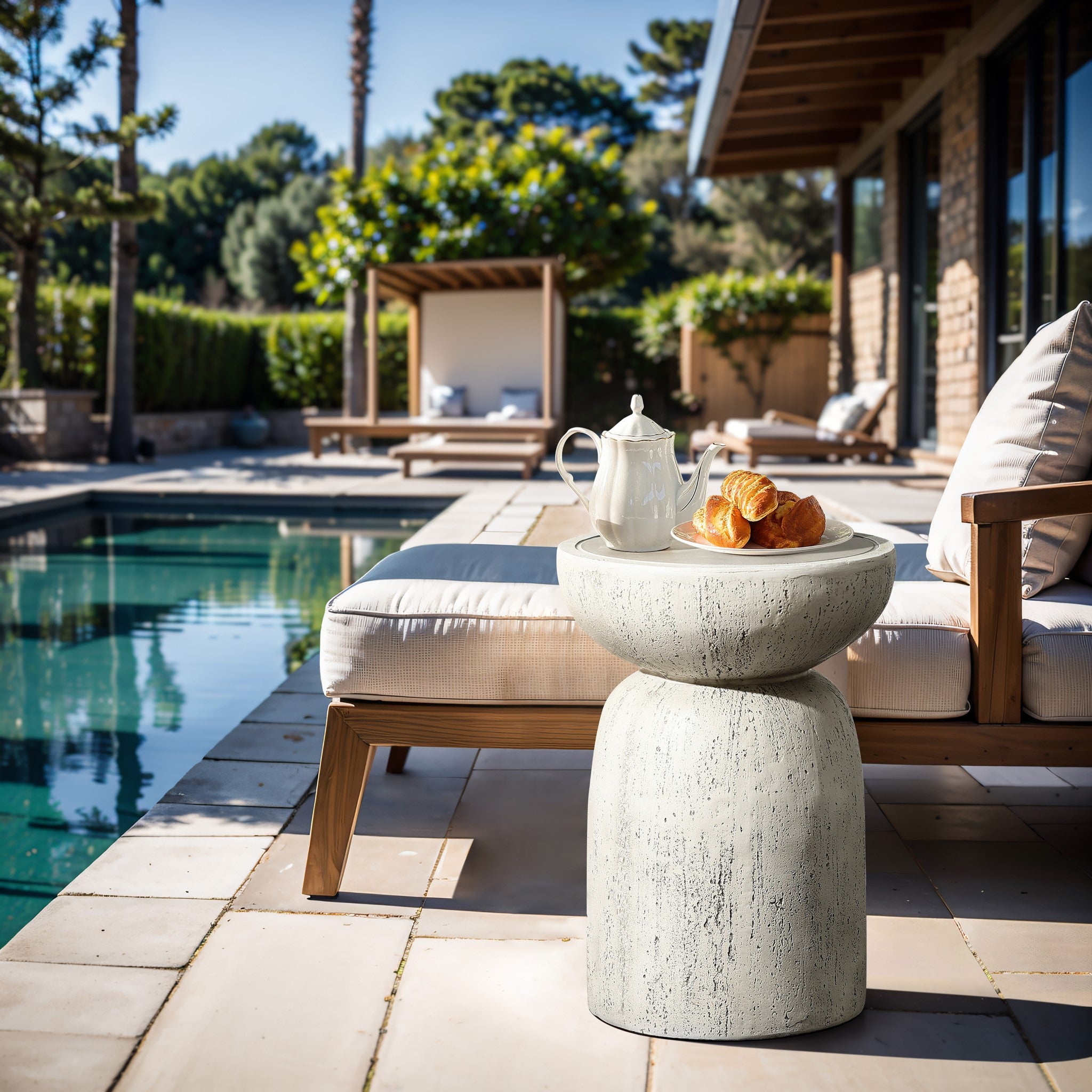 A serene poolside features a lounge chair with cushions and a LuxenHome Rustic off White and gray outdoor side table, 19.7 h, holding a teapot and croissants. The setting is surrounded by lush greenery and a clear blue pool under the sunny sky.