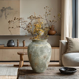 A LuxenHome rustic gold and brown terracotta urn vase (13.6 h) with dried branches adorns a wooden coffee table in a minimalist living room. A beige couch, neutral decor, pottery, and a large abstract painting complete the serene earth-toned setting.