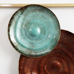 Two ceramic plates hang on a wall as part of an abstract metal wall decor ensemble. The top plate is turquoise with a glossy finish, while the lower plate is dark brown with a matte finish, creating a delightful play of textures in this modern art-inspired display.