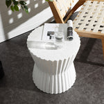 A speckled white fluted hourglass outdoor side table holds a glass of water, an open magazine, and a folded newspaper. It stands next to a woven beige chair on the dark tiled floor. Sunlight casts shadows, with a green potted plant partially visible in the background.