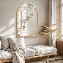 A cozy living room features a light beige sofa and a 34-inch Arch Mirror with scalloped edge framing. A vase of flowers complements the wooden decor, while sunlight streams through the window, casting shadows on the wall.