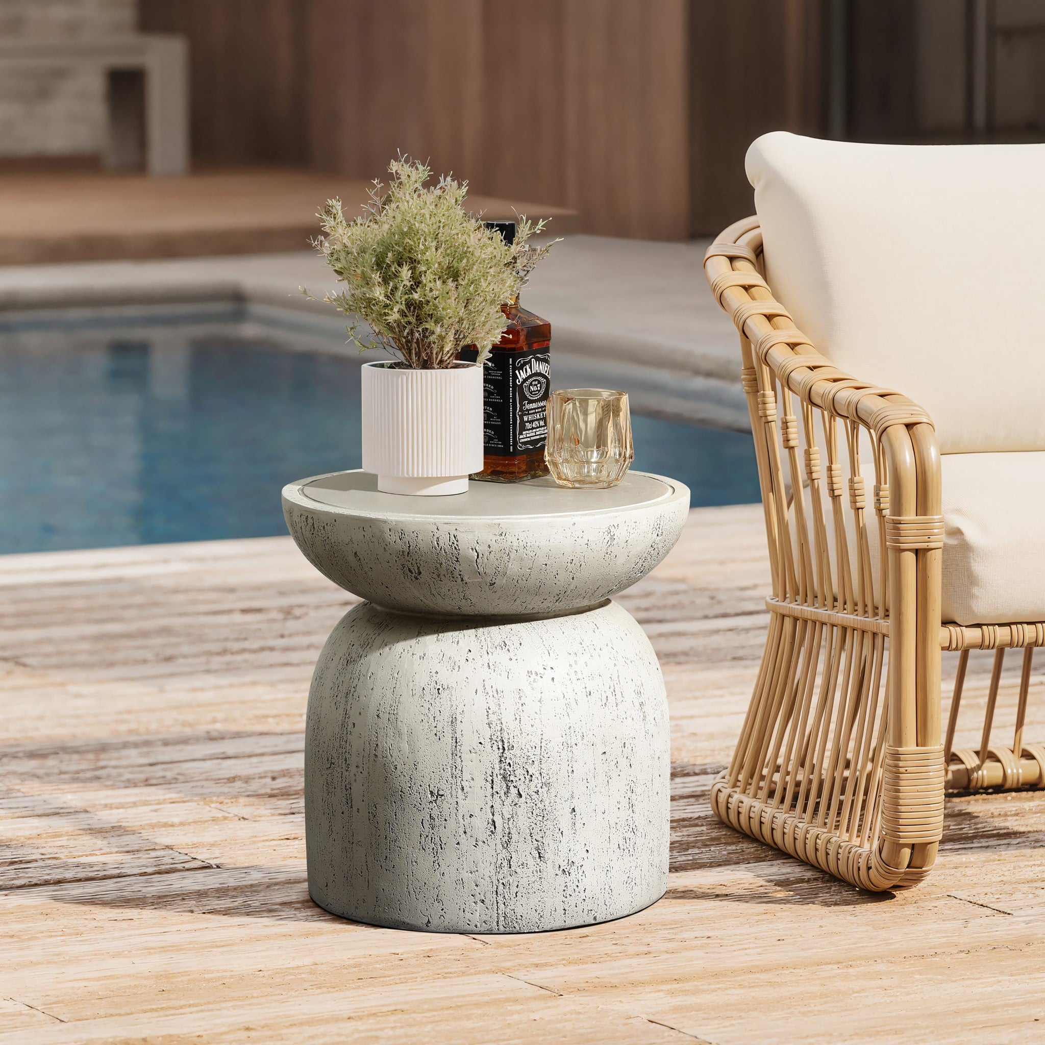 A serene poolside scene features a wicker armchair and a 15.75 high Rustic off White and Gray outdoor side table. The table holds a plant in a white pot, a whiskey bottle, and a glass. In the background, the pools clear water glistens against the wooden decking.