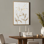 A modern wall decor piece featuring a gold leaf motif hangs above a wooden dining table adorned with three tall candles, two stacked books, and two mugs. To the right, a large vase with branches adds sophistication to this high-end art piece setting.