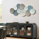 An abstract wall art piece with circular metal discs in various patterns hangs above a dark sideboard with glass doors, showcasing dishes and glassware. A small bowl sits atop the sideboard, adding charm to this vintage metal wall decoration.