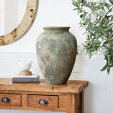 A LuxenHome Rustic gold and brown terracotta urn vase, 13.6 h, sits on a wooden console table with books and a white bird ornament. An olive tree branch and a round mirror enhance the charm, while an antique distressing technique adds timeless elegance to the decor.