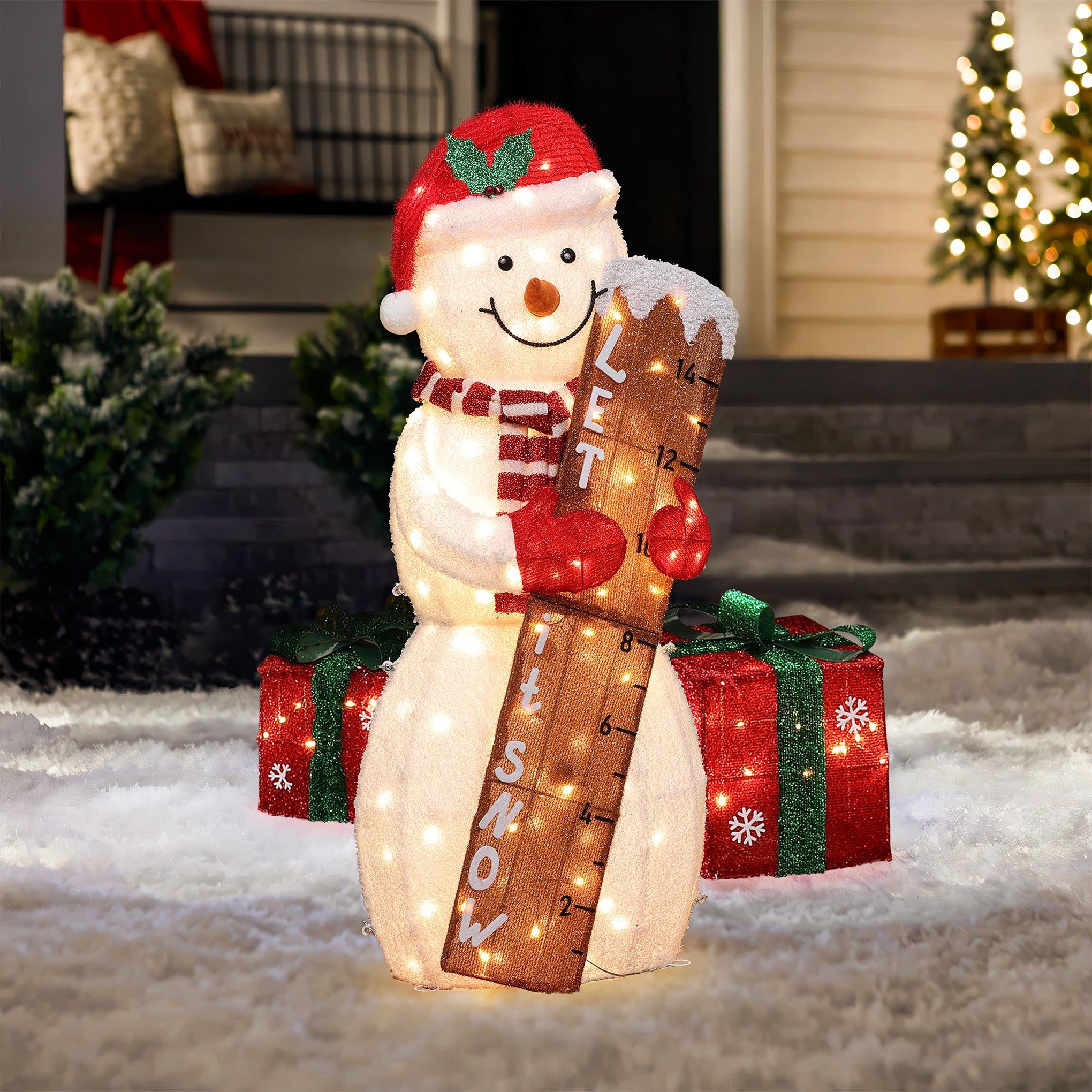 A Snowman Outdoor Holiday Decoration in a Santa hat and scarf holds a ruler marked Let It Snow, surrounded by snow and two large gift boxes in this festive lighted display for charming yard holiday decor at night.