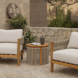 A charming outdoor patio scene featuring two wooden armchairs with white cushions, a round drum-shaped side table crafted from natural acacia wood holding a black pitcher and cups. The setting is enhanced by a stone wall backdrop adorned with lush plants.