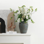 Terracotta vase with white flowers and greenery sits on a white mantel beside books, wooden bird figurines.