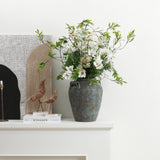 Terracotta vase with white flowers and greenery sits on a white mantel beside books, wooden bird figurines.